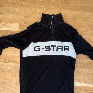 Svart half zip tröja från G-Star - Svart långärmad tröja från G-Star med bred grå panel och stor logga över bröstet. Tröjan har hög krage och half zip-dragkedja framtill. Mjuk och skön känsla, perfekt för en chill och stilren look.