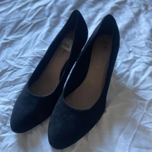 Svarta mockapumps från Tamaris - Klassiska svarta pumps från Tamaris i mjuk mocka. Skorna har spetsig tå och en stabil klack som ger en snygg siluett. Perfekta för dig som gillar stilrena och tidlösa skor med en elegant touch.