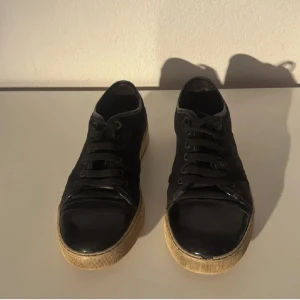 Svarta sneakers från Lanvin - Snygga svarta sneakers från Lanvin med klassisk låg profil. Skorna har svarta snören, rund tå och beige sula med tydlig text undertill. Skorna är andvända och har tydliga defekter men har mycket kvar att ge. Hör av dig vid frågor :)