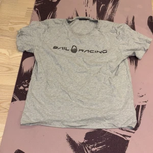 Grå t-shirt från Sail Racing - Snygg grå t-shirt från Sail Racing med svart logga tryckt på bröstet. Klassisk rund halsringning och korta ärmar. Tillverkad i mjuk bomull som känns skön mot huden. Perfekt för en avslappnad och sportig stil.