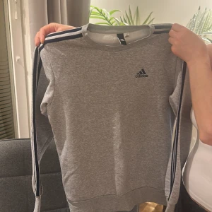 Grå Adidas sweatshirt (priset kan diskuteras vid snabb affär) - Grå sweatshirt från Adidas med klassiska svarta ränder längs ärmarna och Adidas-logga på bröstet. Tröjan har rund halsringning och långa ärmar. Perfekt för en sportig och avslappnad stil. Den är också helt i nyskick och knappt använd!
