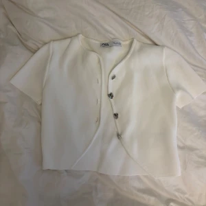 Vit croppad kofta från Zara - Superfin vit croppad kofta från Zara med kort ärm och snygga knappar framtill. Koftan har en enkel, stilren design och är perfekt att slänga över ett linne. Materialet känns mjukt och lätt, vilket gör den skön att bära under varmare dagar.