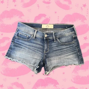 Blå vintage low rise short shorts från Hollister - Snygga blå jeansshorts från Hollister med låg midja och råa, fransiga benslut. Klassisk femficksmodell med slitningar och ljus tvätt. Perfekta för sommardagar och festivaler. Materialet är mjukt och stretchigt med 98% bomull och 2% elastan. Micro mini shorts short shorts low rise lågmidje. 