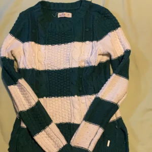 Randig stickad tröja från Hollister XS - Mysig stickad tröja från Hollister i storlek XS. Tröjan har breda gröna och vita ränder, kabelstickat mönster och rund hals. Långärmad och perfekt för kyligare dagar. Klassisk Hollister-logga i metall vid nederkanten.