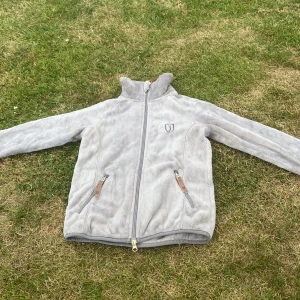 Grå fleecejacka från Jacson 158/164 - Mysig grå fleecejacka från Jacson med hög krage och hel dragkedja. Jackan har två fickor med dragkedja och snygga detaljer i läderimitation. Perfekt för kyliga dagar och enkel att matcha med det mesta. Fläck som vi kommer tvätta bort innan den skickas. 