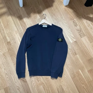 Mörkblå Stone Island sweatshirt - Säljer en mörkblå sweatshirt från Stone Island Junior med klassisk patch på vänster ärm. Tröjan har rund halsringning, ribbade muddar och en clean look som passar till det mesta. Perfekt för dig som gillar streetwear och vill ha något stilrent.