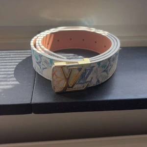 Louis Vuitton vitt bälte med LV-spänne - Snyggt vitt bälte från Louis Vuitton med ikoniskt LV-spänne i guld. Bältet har färgglada detaljer och klassiskt monogrammönster. Perfekt accessoar för att lyfta din outfit med exklusiv känsla.