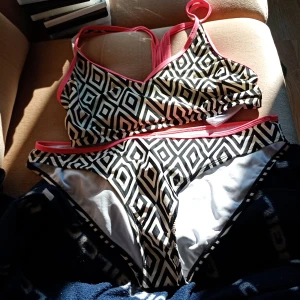 Svartvit bikini med grafiskt mönster - Snygg bikini med svartvitt grafiskt rutmönster och rosa kantband. Överdelen har sportig skärning med breda axelband och trosorna har klassisk passform. Perfekt för stranden eller poolen i sommar!