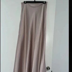 Ljusbeige satinkjol med dragkedja - Supersnygg långkjol i ljusbeige satin-look med lätt glans. Kjolen har en rak och flowy passform och stängs med en diskret dragkedja i sidan. Perfekt för dig som gillar en clean och stilren vibe. Passar till allt från sneakers till boots.