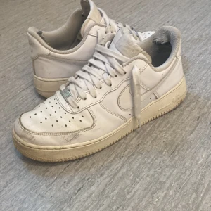 Nike Air Force 1 vita sneakers - Klassiska Nike Air Force 1 sneakers i helvitt skinn med perforerad tå, rund tå och platt sula. Swoosh-logga på sidorna och metallbricka med 'AF1' på snörningen. Perfekta för en clean streetstyle-look.