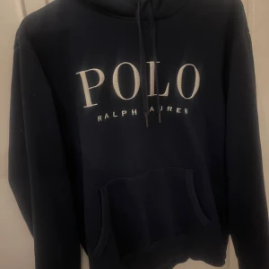 Mörkblå hoodie från Polo Ralph Lauren - Mörkblå hoodie från Polo Ralph Lauren med stor vit logga framtill och broderad polospelare på ärmen. Klassisk känguruficka, snörning i huvan och mjukt material. Perfekt för dig som gillar stilren streetwear med premiumkänsla. Relativt använd men mycket kvar att ge! 