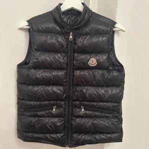 Svart dunväst från Moncler - Snygg svart dunväst från Moncler med klassisk logga på bröstet. Västen har hög krage, två fickor med dragkedja och quiltad design. Perfekt att slänga över en hoodie eller tröja för extra värme.