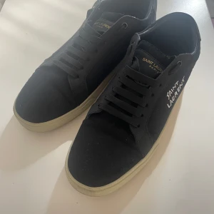 Svarta Saint Laurent sneakers - Svarta sneakers från Saint Laurent med vit logga broderad på sidan och guldtext på plösen. Skorna har svart snörning, rund tå och vit platt sula. Tillverkade i canvas med klassisk, stilren design som passar till det mesta. Säljer då de är för små, har inklusive dust bag