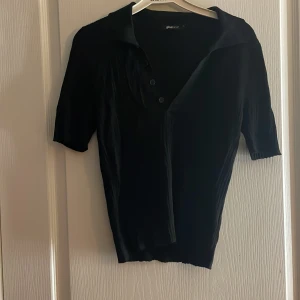 Svart pikétröja från Gina Tricot - Svart pikétröja från Gina Tricot i storlek L. Tröjan har klassisk krage, knäppning med tre knappar och korta ärmar. Materialet är lätt ribbat och ger en snygg struktur. Perfekt till jeans eller kjol för en clean look.