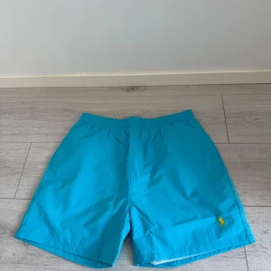 Turkosa badshorts Polo Ralph Lauren  - Turkosa badshorts från Polo Ralph Lauren i storlek XL sitter dock med som L. Klassisk modell med elastisk midja, innerbyxa i mesh och bakficka. Gult broderat logotyp på benet och gul insida i midjan. Perfekta för stranden eller poolen.