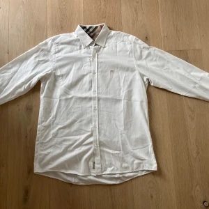 Vit skjorta från Burberry med rutigt foder - Klassisk vit skjorta från Burberry med lång ärm och knappar framtill. Kragen har det ikoniska rutiga Burberry-mönstret på insidan och bröstet pryds av en broderad logga i beige. Skjortan är i bomull och har en normal passform, perfekt för en clean och stilren look.