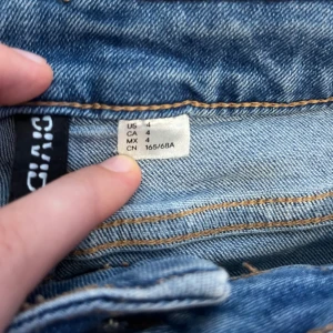 Blå bootcut jeans från Divided - Snygga blå jeans från Divided med klassisk bootcut-modell och normal midja. Jeansen har fem fickor, dragkedja och knapp framtill samt coola slitningar för en avslappnad look. Perfekta för dig som gillar retrovibbar och vill ha ett par jeans som sticker ut.