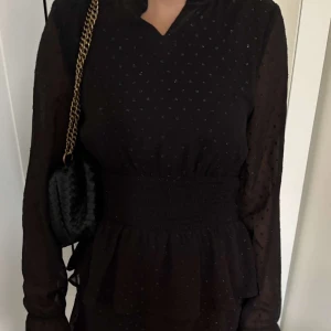 Svart kort prickig klänning - Supersöt svart kort klänning med små prickar, transparenta långärmar och markerad midja med smock. Volangdetaljer nertill ger en flowy känsla. Perfekt för dig som gillar en feminin och trendig look.