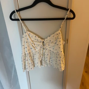 Söt blommig linnetopp med snörning - Ljus linnetopp med blått blommönster och smala axelband. Toppen har en söt volangkant nertill och snörning framtill som ger en fin detalj. Perfekt för varma dagar och somriga outfits.