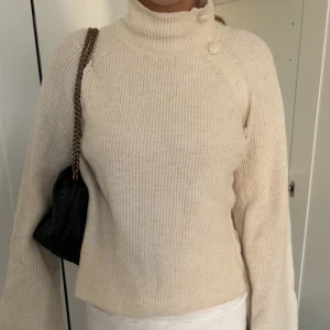 Beige ribbstickad polotröja med knappar - Supermysig beige ribbstickad polotröja med höga krage och snygga knappar på axeln. Tröjan har lång ärm och en relaxed fit som är perfekt för lager-på-lager. Enkel att matcha med både jeans och kjol för en chill look. (En knapp har trillat av, men fortfarande i bra skick!)