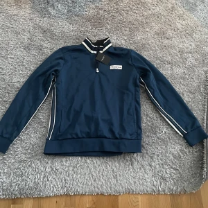 Blå half zip tröja från Björn Borg - Snygg marinblå tröja från Björn Borg med half zip och vita ränder på kragen och längs ärmarna. Tröjan har lång ärm, ribbade muddar och en broderad logga på bröstet. Perfekt för en sportig och stilren look. Tröjan är helt oanvänd och har prislappen kvar