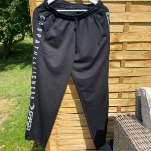 Svarta gymbyxor från Gasp - Svarta gymbyxor från Gasp med tryckt logga och texten 'Gasp Athletic Wear' längs ena benet. Byxorna har resår och snörning i midjan samt raka ben. Perfekta för gymmet eller chill hemma. Materialet är mjukt och bekvämt. Har en defekt på ena benets tryck där det går som ett svart veck genom gasp trycket. Vid frågor eller funderingar skicka ett meddelande💫