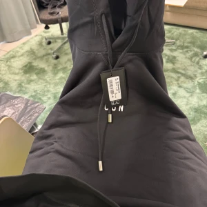 Svart hoodie DSQUARED2 ICON M - Svart hoodie från DSQUARED2 med vit 'ICON'-logga på bröstet. Klassisk modell med huva och snörning, samt ribbade muddar. Tillverkad i mjuk bomull för en skön känsla. Perfekt för dig som gillar streetwear och vill sticka ut med en ikonisk look.