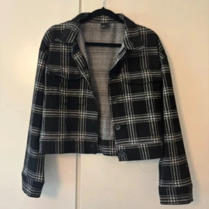 Rutig overshirt från Gina Tricot XS - Snygg croppad overshirt från Gina Tricot i svart och vitt rutigt mönster. Jackan har klassisk krage, knappar framtill och två bröstfickor. Perfekt för lager-på-lager och ger en cool vibe till din outfit.