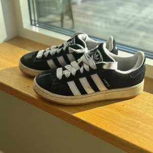 Adidas Campus svarta sneakers  - Svarta Adidas Campus sneakers i mocka med klassiska vita ränder och vit logga på plösen. Skorna har vit sula och vita skosnören för en clean och tidlös look. Perfekta för dig som gillar streetstyle och vill ha en ikonisk sneaker i garderoben.