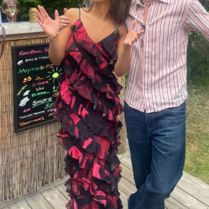 Lång röd och svart volangklänning - Säljer en maxiklänning med tunna axelband i rött och svart med volanger över hela klänningen. Klänningen har ett blommigt mönster och är i ett lätt, flowy material som känns perfekt för sommarens alla tillfällen.