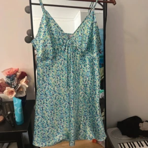 Blommig sommarklänning med knyt - Söt kort blommig klänning i blått och grönt med smala axelband och v-ringning. Klänningen har knyt framtill och är perfekt för varma dagar. Lätt och flowy känsla, passar dig som gillar en avslappnad men ändå söt stil.