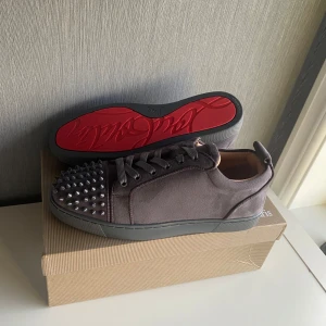 Christian Louboutin sneakers med nitar - Snygga mörkgrå sneakers från Christian Louboutin med klassisk röd sula och coola silverfärgade nitar på tån. Skorna har snörning och är tillverkade i mocka med läderdetaljer. Perfekt för dig som vill sticka ut med exklusiv streetstyle.