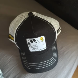 Snoopy keps från Capslab Peanuts - Svart och vit keps från Capslab med mesh på sidorna och bak, samt broderad patch framtill med Snoopy och Woodstock från Peanuts. Snygga kontrastsömmar och detaljer med Peanuts-logga på sidan. Perfekt för dig som gillar coola accessoarer och serietidningsstil.