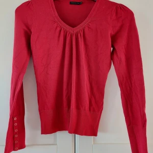Rubinröd v-ringad tröja från Stockh LM - Djupt röd v-ringad tröja från Stockh LM i mjuk bomullsmix. Långa ärmar med snygga knappar längs mudden och lätt puff vid axlarna. Perfekt för dig som gillar stilrena detaljer och en feminin touch.