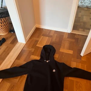 Svart Moncler hoodie med logga - Svart hoodie från Moncler med klassisk broderad logga på bröstet. Tröjan har huva med snörning och långa ärmar. Tillverkad i mjuk bomull som känns skön mot huden. Perfekt för dig som gillar stilrena och exklusiva plagg.