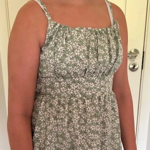 Grön blommig sommarklänning - Söt ljusgrön kortklänning med vita blommor och smala axelband. Klänningen har rynkade detaljer upptill och smockad rygg för en bekväm passform. Perfekt för varma dagar och har en luftig känsla.