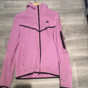 Ljuslila Nike tech jacka  - Nike hoodiejacka i ljuslila med svarta detaljer och dragkedja hela vägen. Jackan har huva, fickor på sidorna och en extra ficka med dragkedja på ena ärmen. Den har en synlig defekt på bakre armen, se bild. Storlek M
