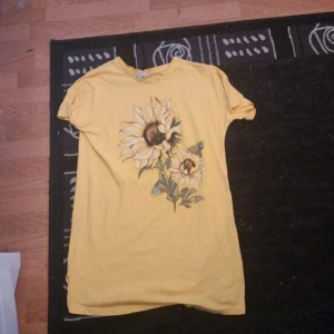 Gul t-shirt med solrosmotiv - Gul t-shirt med korta ärmar och stort tryck av solrosor på framsidan. Skön och avslappnad passform, perfekt för dig som gillar färg och blommiga prints. Tillverkad i mjuk bomull som känns nice mot huden.