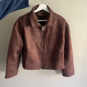 Brun mockajacka från Emma Lisa Stockholm - Säljer en brun mockajacka från Emma Lisa Stockholm. Jackan har en croppad passform, dragkedja framtill och klassisk krage. Perfekt för dig som vill ha en stilren och trendig jacka i garderoben. Den har aldrig använts så den är helt ny.  