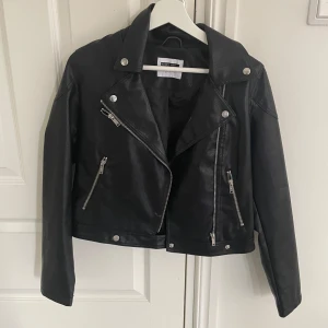 Svart skinnjacka från Noisy May - Säljer en svart skinnjacka från Noisy May med klassisk bikerstil. Jackan har silvriga dragkedjor, knappar och detaljer samt snedställd dragkedja framtill. Perfekt för en cool och stilren look.