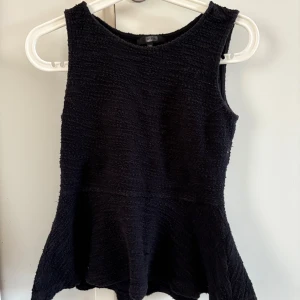 Svart ärmlös topp från Bik Bok - Säljer en svart ärmlös topp från Bik Bok i storlek XS. Toppen har en lätt utställd peplumform och är tillverkad i ett strukturerat tyg som ger en snygg look. Perfekt att styla med jeans eller kjol! Inga defekter