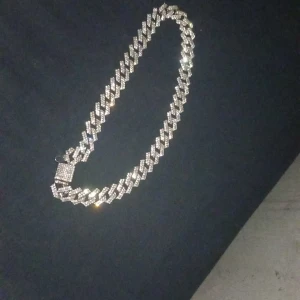 Glittrigt silverfärgat halsband - Säljer ett chunky halsband i silverfärg med glittrande stenar. Kedjan har ett brett, platt mönster och ett fyrkantigt lås dekorerat med stenar. Perfekt för att ge din outfit extra bling.