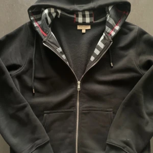 Svart hoodie från Burberry med dragkedja - Säljer en svart hoodie från Burberry i storlek L.  Tröjan har dragkedja, huva med klassiskt rutigt foder och broderad logga på bröstet. Tillverkad i mjuk bomull och har fickor framtill. Perfekt för en stilren och bekväm look. Storleken känns mer Som L ej Xl