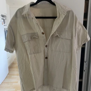 Beige kortärmad skjorta med fickor - Säljer en beige kortärmad skjorta med två bröstfickor och knappar framtill. Skjortan har en avslappnad passform och är perfekt för varma dagar. Enkel och stilren design som passar till mycket.
