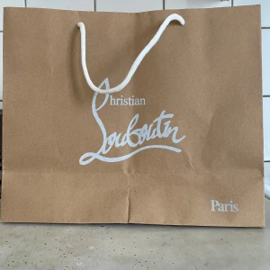 Christian Louboutin shoppingpåse - Snygg brun shoppingpåse från Christian Louboutin med vit logotyp och texten Paris. Påsen har ett vitt snöre som handtag och är perfekt för att bära skor eller accessoarer.