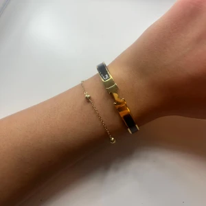 Svart och guld armband - Svart armband med guld detaljer. Armbandet har en H-loggan framtill och är stelt i modellen. Aldrig använd.