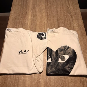Vit t-shirt från Comme des Garçons Play - Säljer två vita t-shirts från Comme des Garçons Play med det ikoniska hjärtmotivet och logga på bröstet. En har stort svart hjärta med ögon, den andra har liten logga och hjärtdetalj på ärmen. Perfekt för en stilren och trendig look.                           OBS:1 FÖR 399 2 FÖR 799