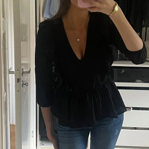 Svart blus med volang och v-ringning - Säljer en svart blus med v-ringning och volangdetaljer längs axlarna och framtill. Blusen har trekvartsärm och är tillverkad i ett lätt, något transparent material med struktur. Perfekt för dig som vill ha en stilren men ändå trendig topp.