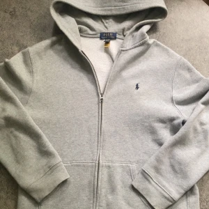 Grå hoodie från Polo Ralph Lauren - Säljer en klassisk grå hoodie från Polo Ralph Lauren i storlek XL. Tröjan har dragkedja, huva och den ikoniska broderade loggan på bröstet. Perfekt för en avslappnad stil och enkel att matcha med det mesta. Skriv vid minsta lilla fundering! 🤗