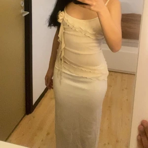 Champagnefärgad topp med volangdetaljer - Säljer en elegant champagnefärgad topp med smala axelband och volangdetaljer längs överdelen och midjan. Toppen har en dekorativ blomdetalj på axeln och en figurnära passform som framhäver siluetten.